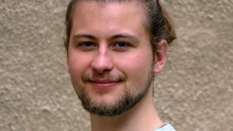 Portrait of a smiling man with a light beard and hair tied back, wearing a light blue shirt, standing against a beige textured wall.