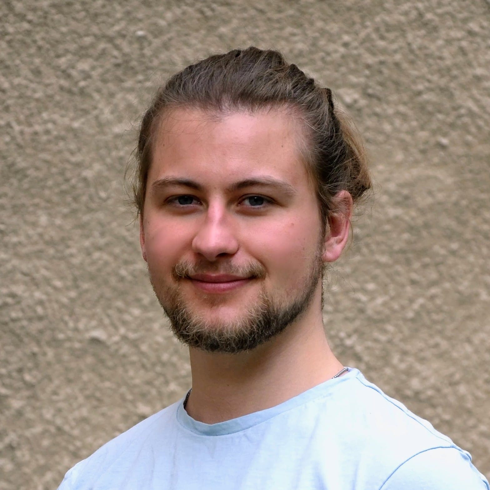 20260506_0002~2 Portrait of a smiling man with a light beard and hair tied back, wearing a light blue shirt, standing against a beige textured wall.