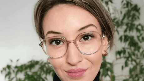Woman with short hair and round glasses, wearing a black turtleneck, smiling softly indoors with green plants behind her