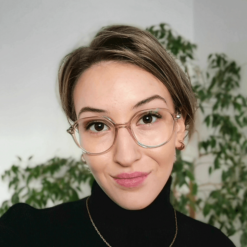 Kwadrat na strone (7) Woman with short hair and round glasses, wearing a black turtleneck, smiling softly indoors with green plants behind her