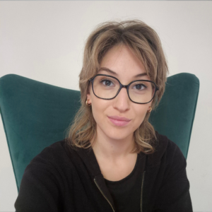 Woman with short blond hair and black-rimmed glasses, seated in a teal chair and looking at the camera.