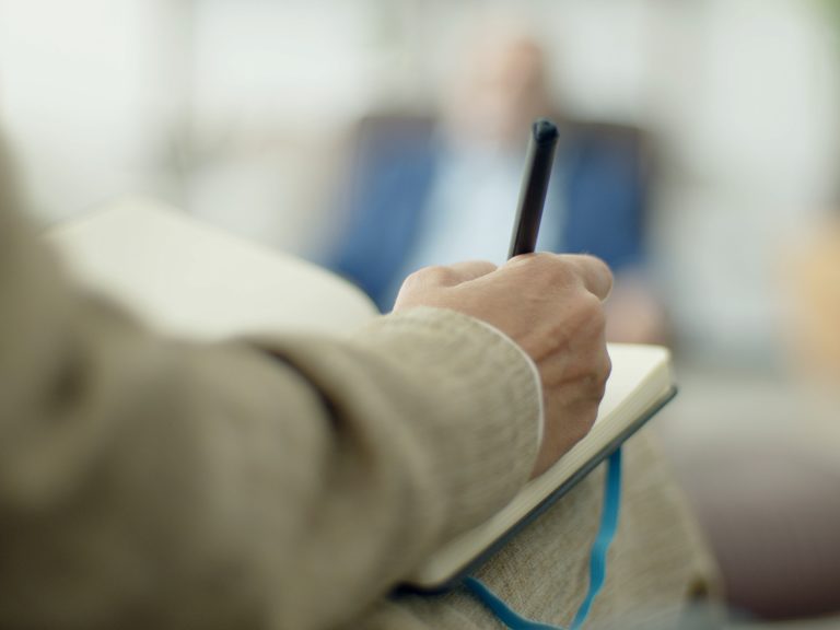 Close-up of a hand holding a pen over an open notebook, with a blurred office background behind.
